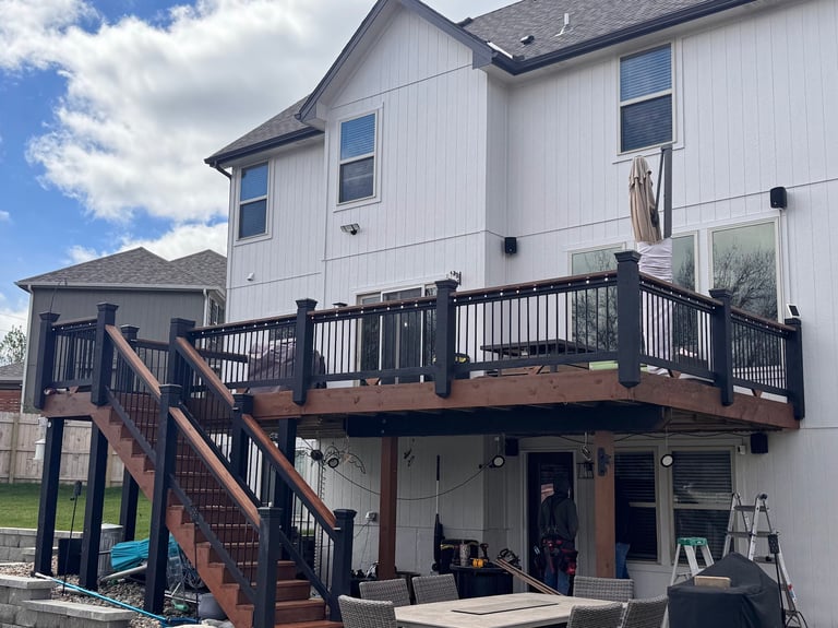 Two-story white house with dark wooden deck, black railings, and stairs under blue sky with clouds