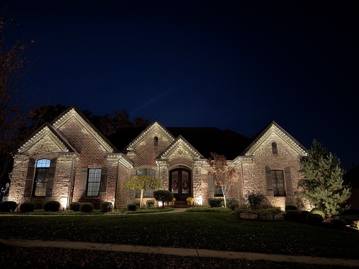 Stone house illuminated at night with landscape lighting, manicured lawn, and wooded backdrop