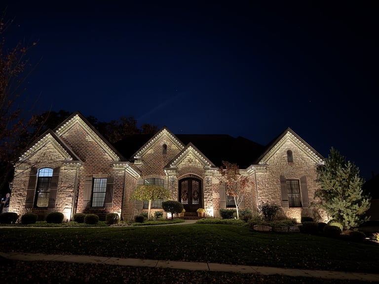 Stone house illuminated at night with landscape lighting, manicured lawn, and wooded backdrop against dark blue sky