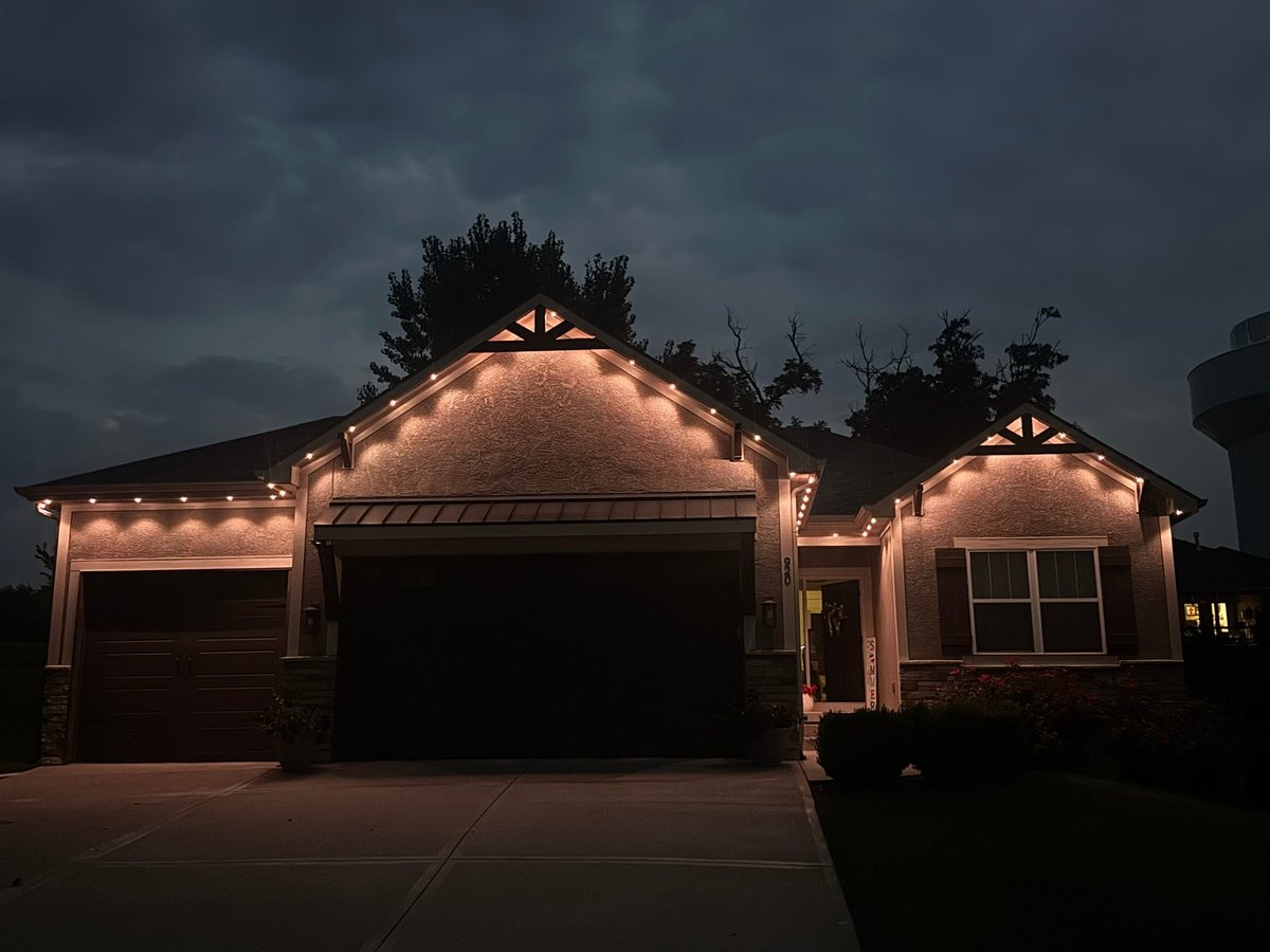 Two-story house illuminated with warm lights at dusk, featuring a large garage and curved roofline against a dark stormy sky