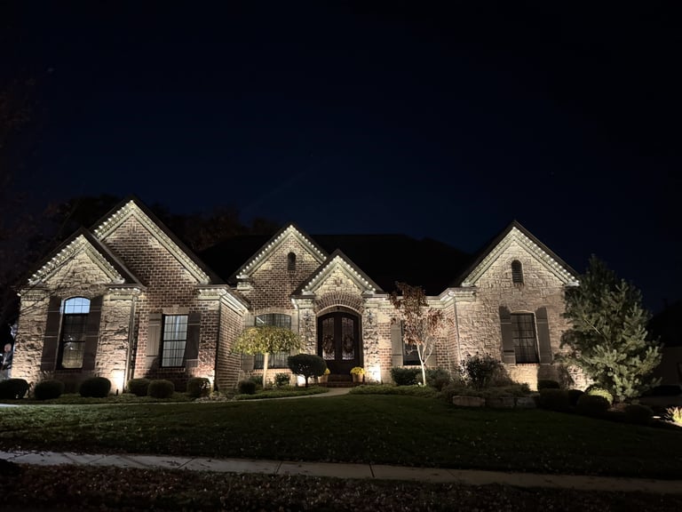 Illuminated stone residential home at night with peaked roofs, landscaping, and warm exterior lighting against dark sky
