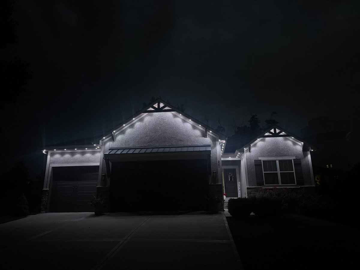 A modern house with architectural details lit by spotlights at night, featuring a prominent garage and dark surroundings