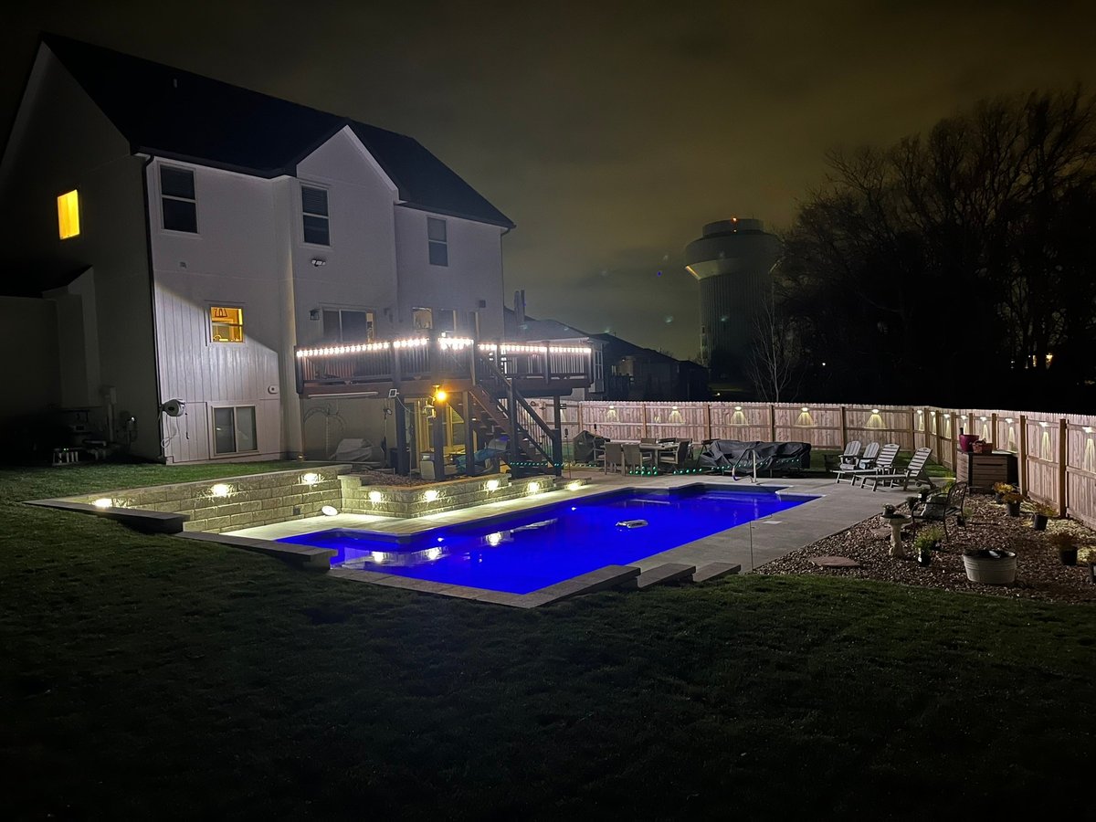 Modern two-story house with illuminated pool at night, white fencing, and landscaped yard with outdoor furniture