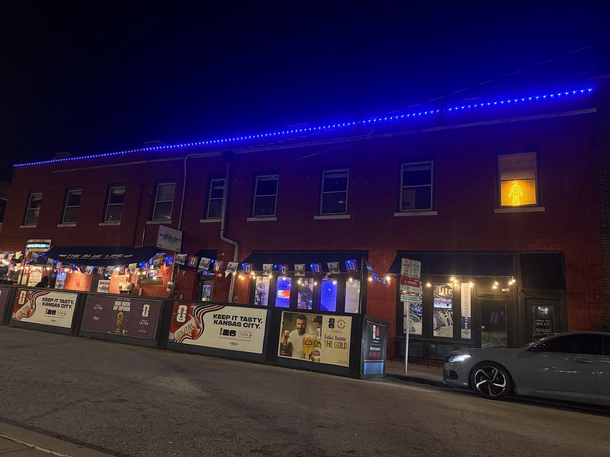 Brick storefront at night with blue neon lighting on roof, illuminated display windows, and parked car outside